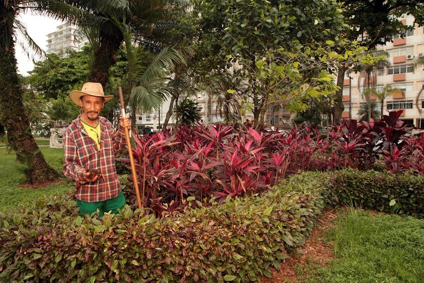 Juvenaldo posa para foto em meio a vegetação no jardim. ele usa chapéu e segura um instrumento de cabo longo. #paratodosverem