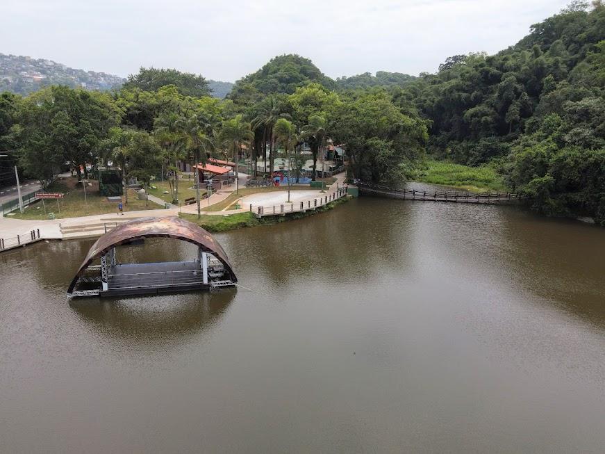 vista aérea da lagoa da saudade, com palco flutuante na água, vegetação e área de lazer ao fundo. #paratodosverem