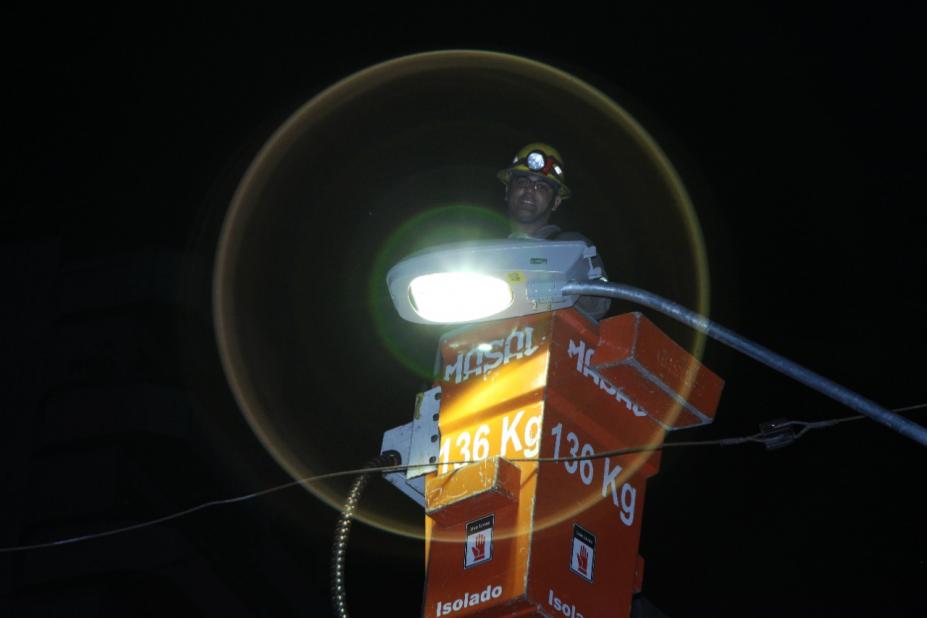 homem está em cesto no alto de poste mexendo em luminária acesa. #paratodosverem