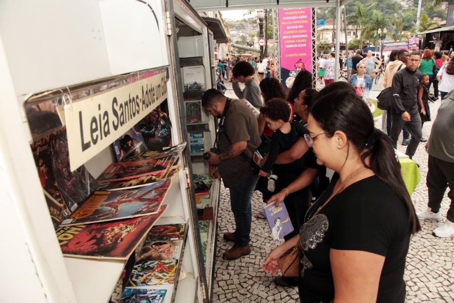pessoas estão em torno de estate de livros montada em praça. #paratodosverem 