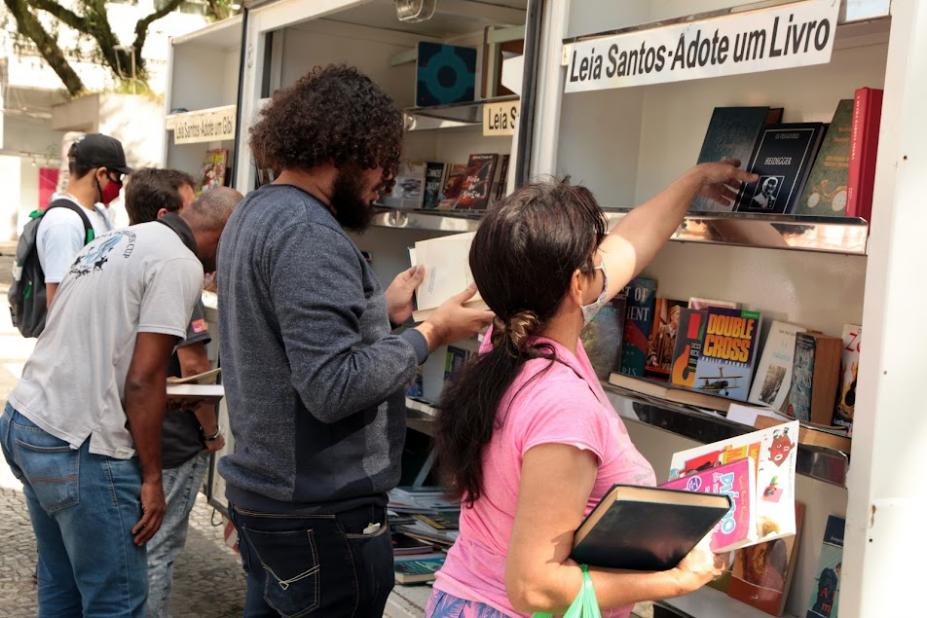 pelo menos cinco pessoas estão diante de estante de bibliioteca na rua. Duas, em primeiro plano, mexem nas publicações. #paratodosverem