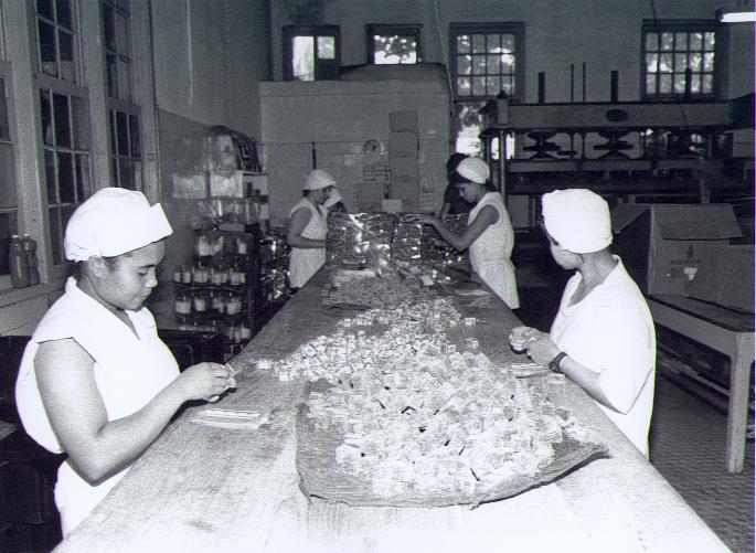 mulheres embrulham balas sobre balcão. Foto antiga em preto e branco. #paratodosverem