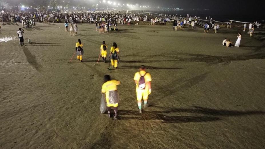 pessoas uniformizadas varrem e coletam lixo na faixa de areia à noite. #paratodosverem 