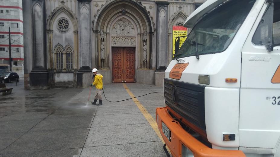 frente de caminhão de lado em primeiro plano e homem esguichando água no pixo ao fundo. Ele está à frente do portal de uma igreja. #paratodosverem