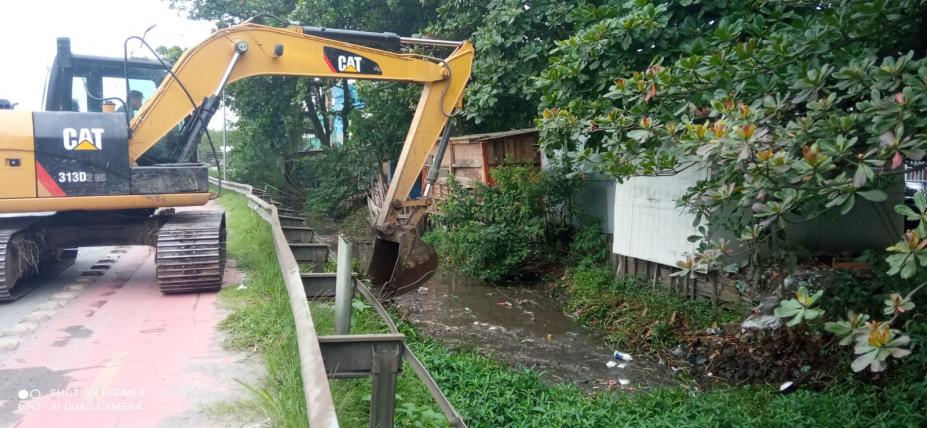 Trator com escavadeira realiza limpeza em córrego ao lado de área verde #paratodosverem