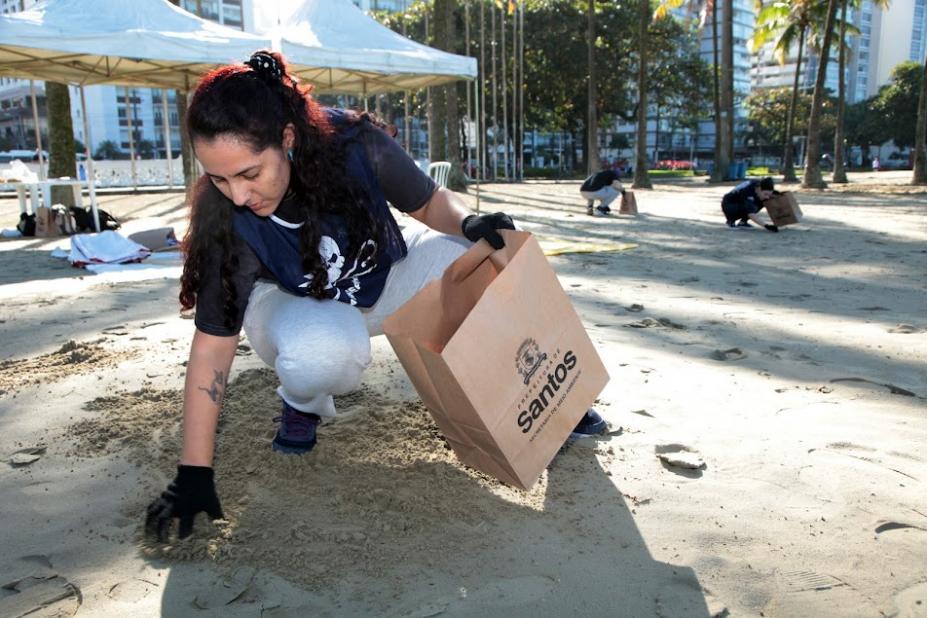 mulher agachada na areia usa luva para recolher lixo e depositá-lo num saco de papel. Ao fundo, há outras pessoas agachadas recolhendo o lixo. #paratodosverem