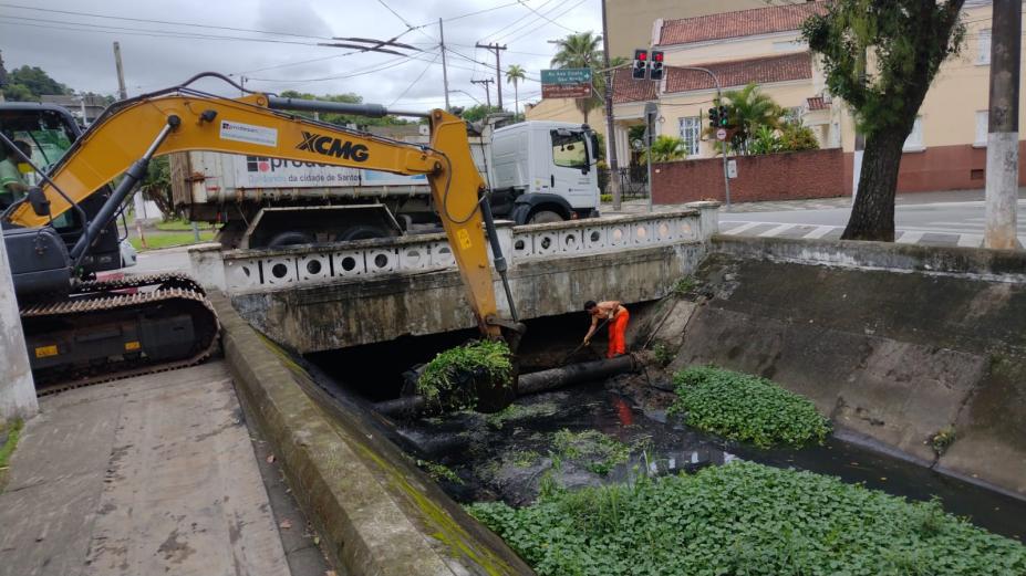 escavadeira retira mato da superfície de canal. Homem está sobre um cano dentro do canal, fazendo a retirada manual de resíduos. #paratodosverem