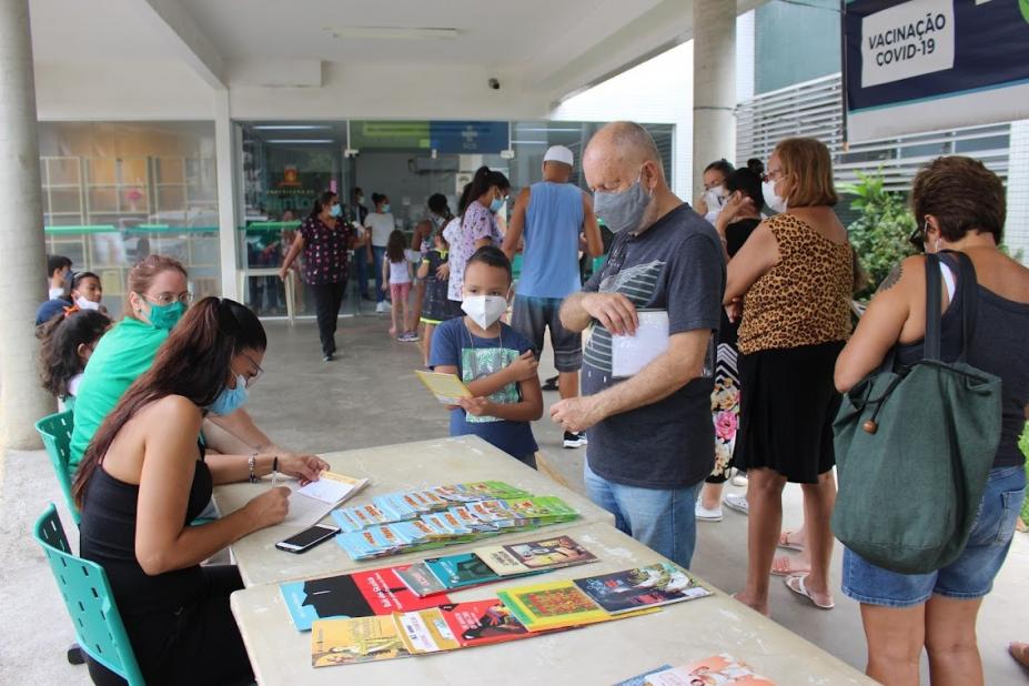 livros expostos sobre uma mesa, com duas pessoas atendendo. Uma criança olha e segura um livro ao lado de um adulto. Há uma filha de  pessoas para vacinação atrás deles. #paratodosverem