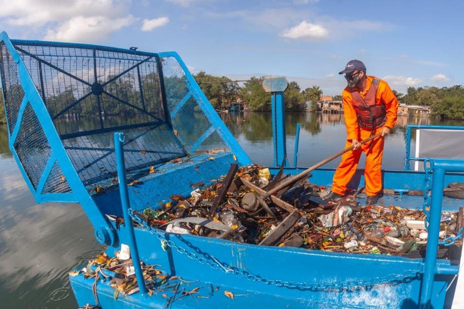 Homem mexe no lixo em uma balsa em cima de um rio #paratodosverem