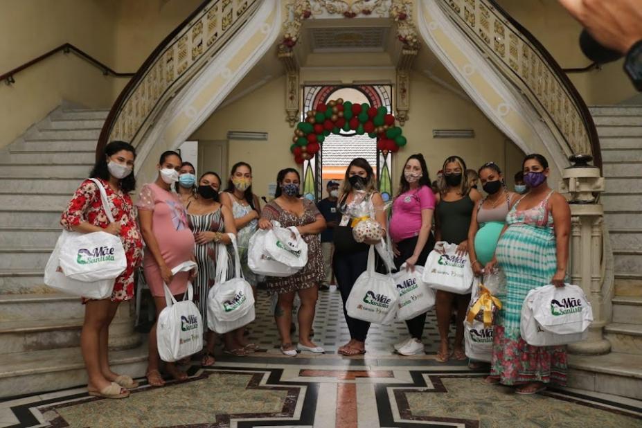 mulheres posam para foto segurando bolsas do programa. Muitas deles estão grávidas. Elas estão diante de duas grandes escadarias no fundo social. #paratodosverem