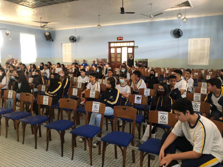 alunos sentados em auditório de escola. Há ventiladores no teto. #paratodosverem