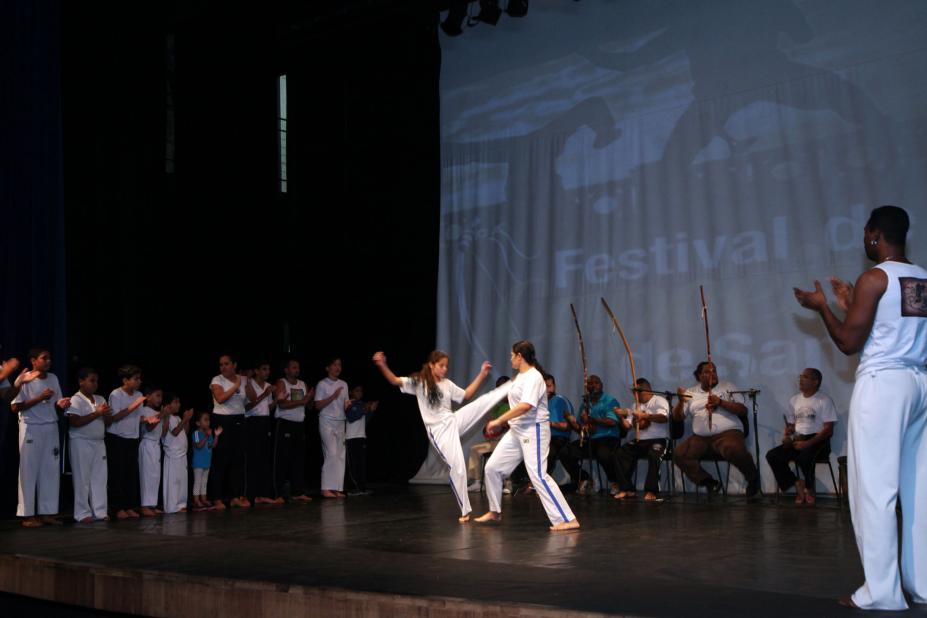 Apresentação de capoeira em palco. Jovens estão ao centro de uma roda jogando a modalidade. Ao redor, outros integrantes do grupo tocam instrumentos típicos