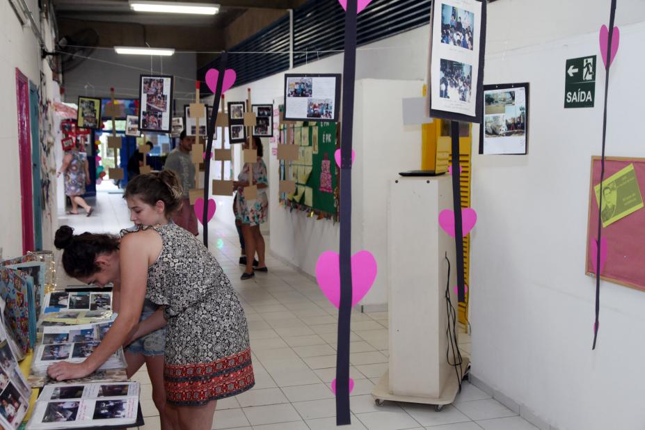 Corredor da escola está decorado com exposição de fotos e corações cor de rosa. À esquerda da imagem há uma mulher e uma criança olhando fotos que estão sobre uma mesa. #Pracegover