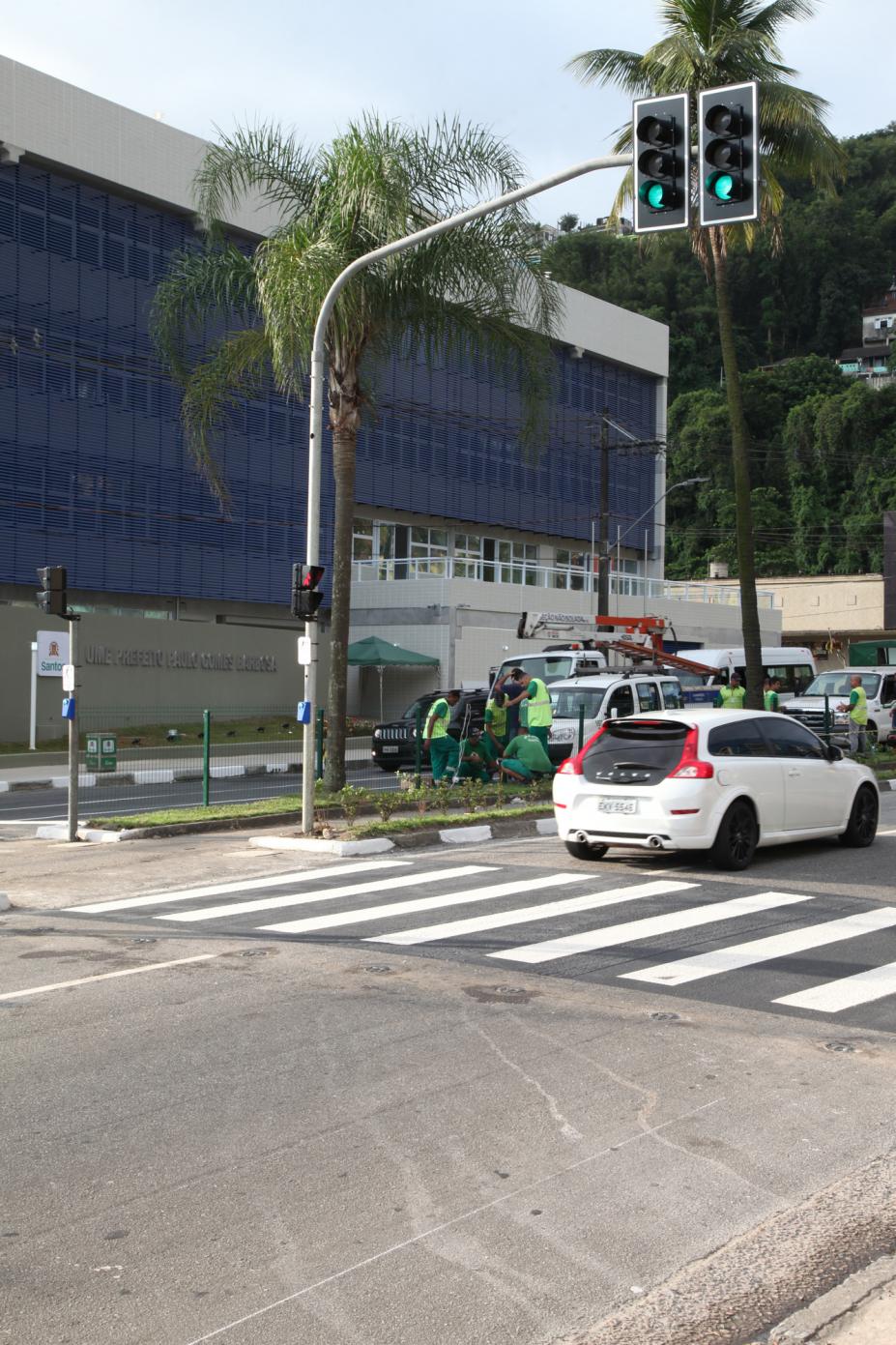 Semáforo em funcionamento em via. Há dois carros passando na pista sobre uma faixa de pedestres. #Pracegover