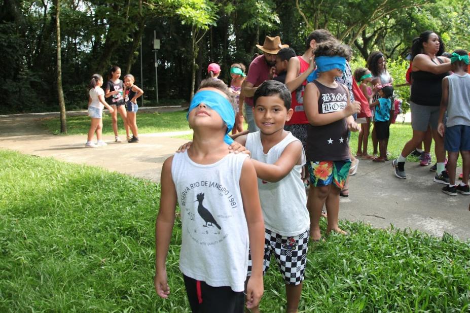 criança empurra outra com os olhos vendados e várias crianças atrás #paratodosverem