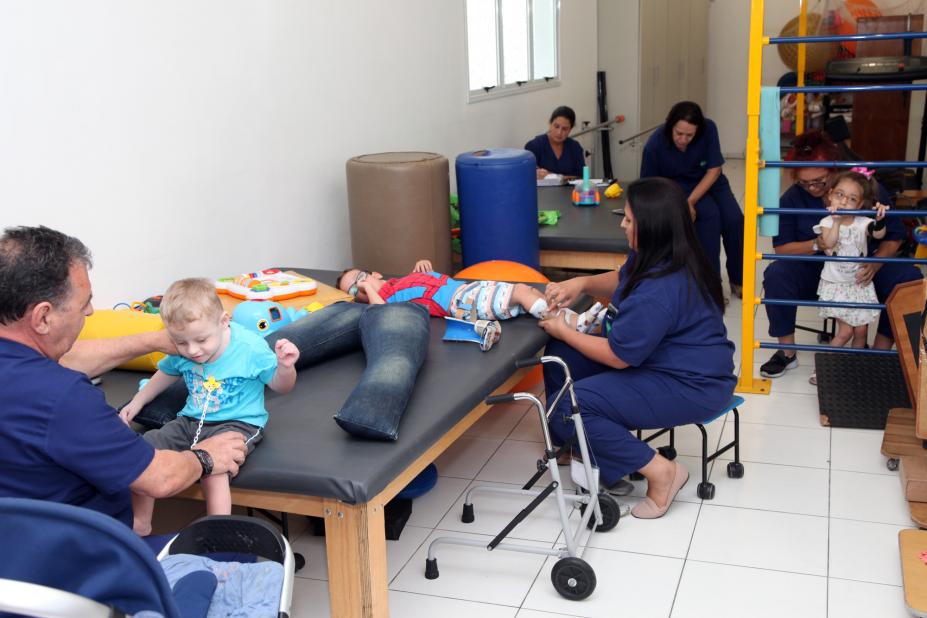 Sala de tratamento fisioterápico, com aparelhos para exercícios. Adultos ajudam crianças a se movimentar. #Pracegover