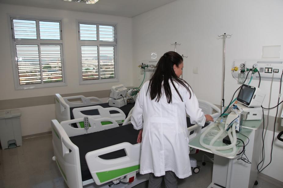 Dois leitos de hospital com equipamentos ao lado. Há uma mulher, de costas para a imagem, mexendo em equipamentos. #Paratodosverem