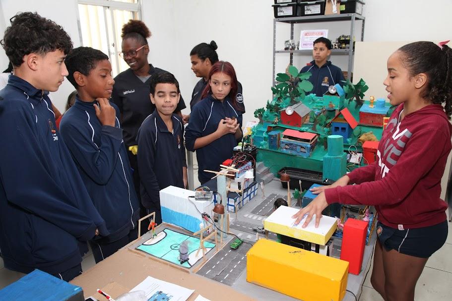 aluna está explicando maquete montada sobre mesa para outros estudantes. todos estão uniformizados. #paratodosverem 