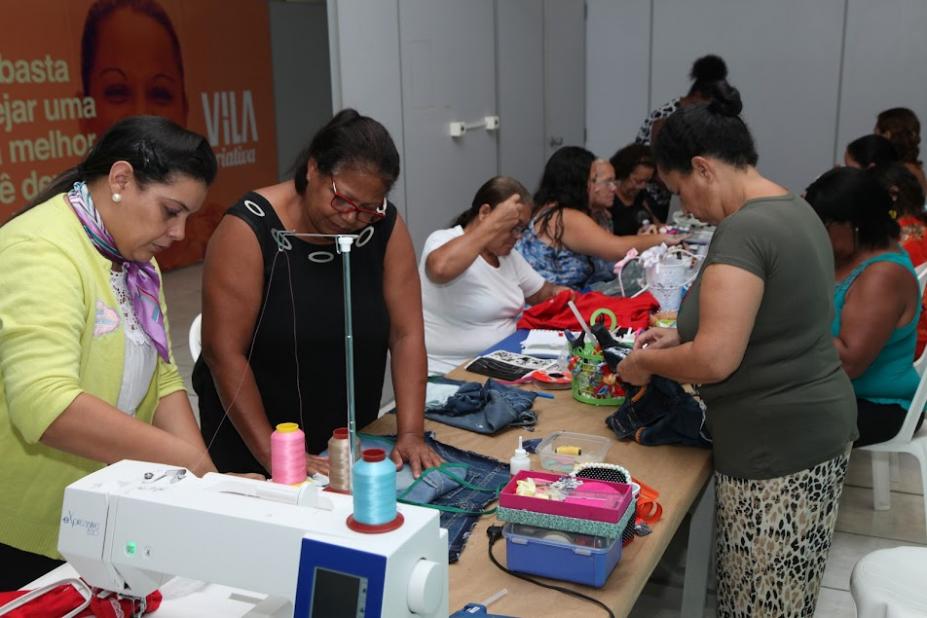 mulheres estão trabalhando em costura em torno de uma mesa. Em primeiro plano está uma máquina de costura. #paratodosverem