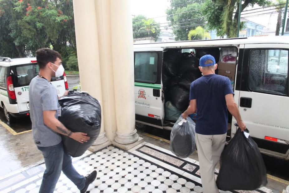 Dois homens carregam sacos com doações em direção a veículo. #paratodosverem