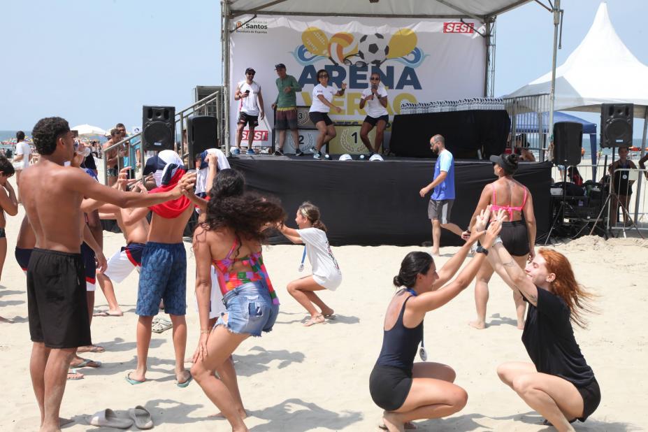 pessoas dançam e se movimentam na faixa de areia diante de um palco em cujo fundo se lê Arena. #paratodosverem 