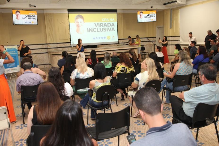 salão com várias pessoas sentadas assistindo a uma apresentação. Ao fundo, em um telão, se lê Virada Inclusiva. #paratodosverem