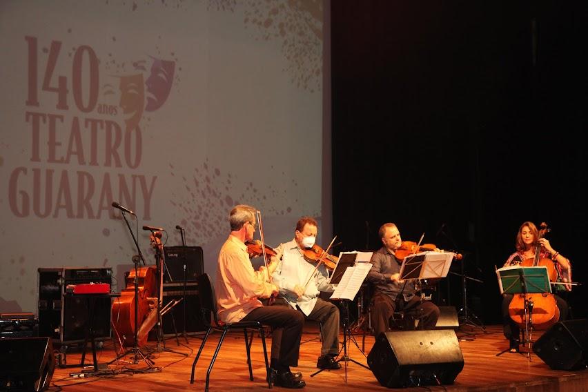 quatro músicos de cordas estão sentados se apresentado num palco. Ao fundo se lê 140 anos Teatro Guarany. #paratodosverem 