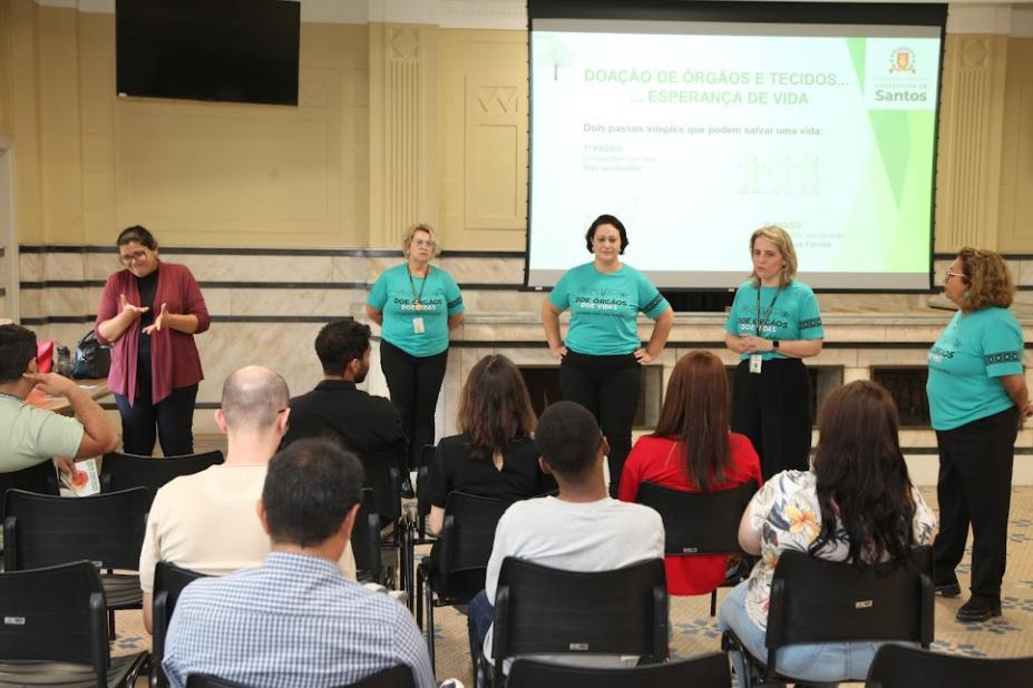 pessoas estão sentadas assistindo palestra de mulheres que estão em pé. Ao fundo um telão com dizeres sobre o transplante de órgãos e tecidos. #paratodosverem 