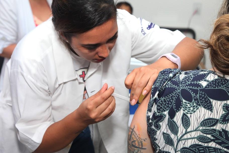 mulher usando avental branco vacina braço esquerdo de mulher que está sentada e de costas para a foto. #paratodosverem 