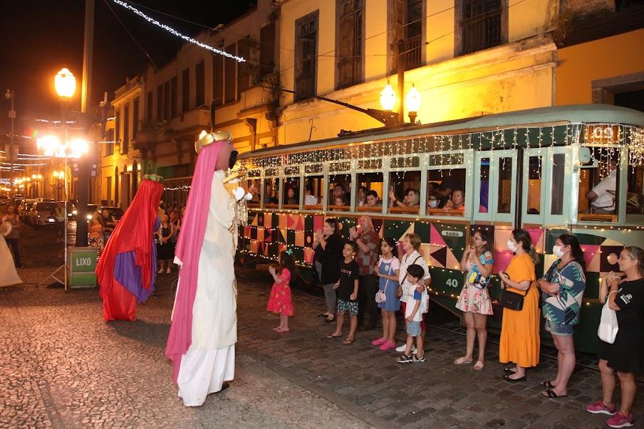 personagens vestidos para interpretar presépio se apresentam diante de bonde cheio de passageiros. #paratodosverem