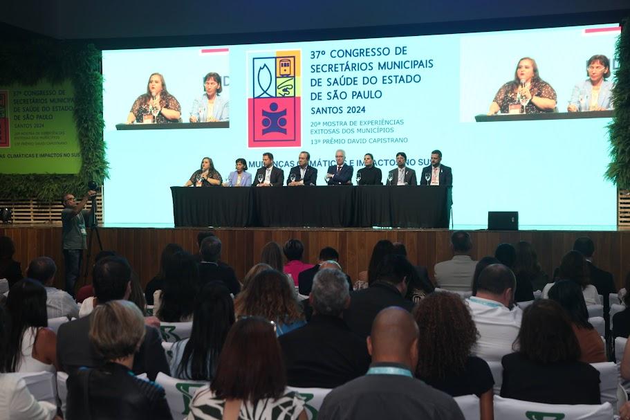 palco com mesa e pessoas sentadas. Ao fundo um telão com imagens. Público está sentado, em primeiro plano, assistindo às apresentações. #paratodosverem 