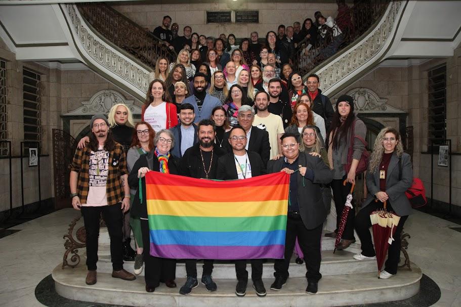 integrantes de conselho posam para foto na escadaria do saguão da prefeitura. à frente, a bandeira lgbt.#paratodosverem