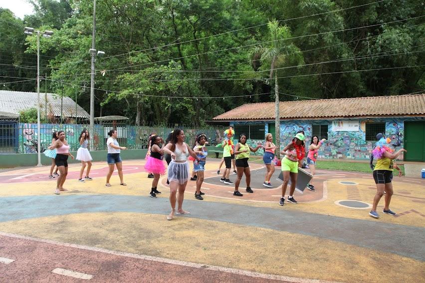 mulheres estão dançando em quadra com grande vegetação ao fundo. #paratodosverem