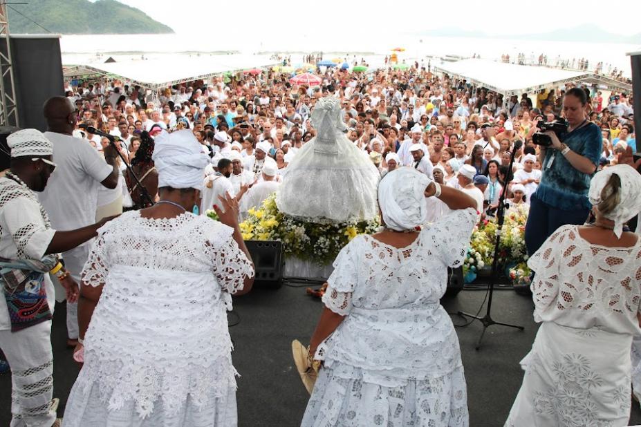 mulheres vestidas de branco estão em palco de costas para a foto. Imagem de iemanjá está à frente com o público ao fundo. #paratodosverem