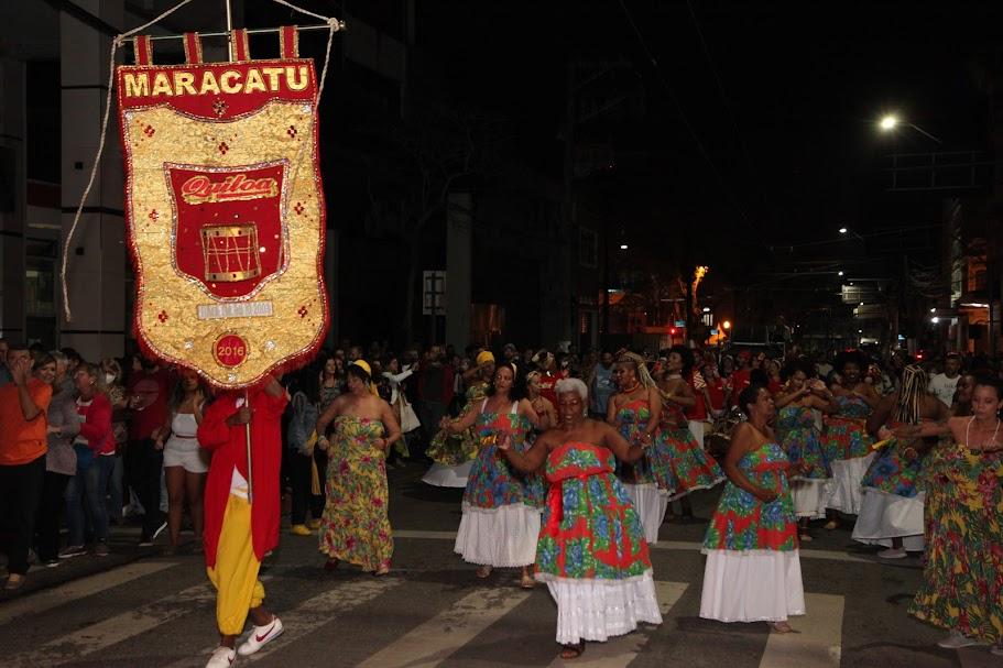 cortejo do grupo maracatu quiloa por rua. Homem à frente segura estandarte e mulheres vem dançado com vestidos coloridos. #paratodosverem