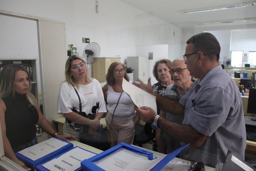 pessoas estão reunidas em um escritório. Um deles mexe em um papel e mostra para os outros. #paratodosverem 