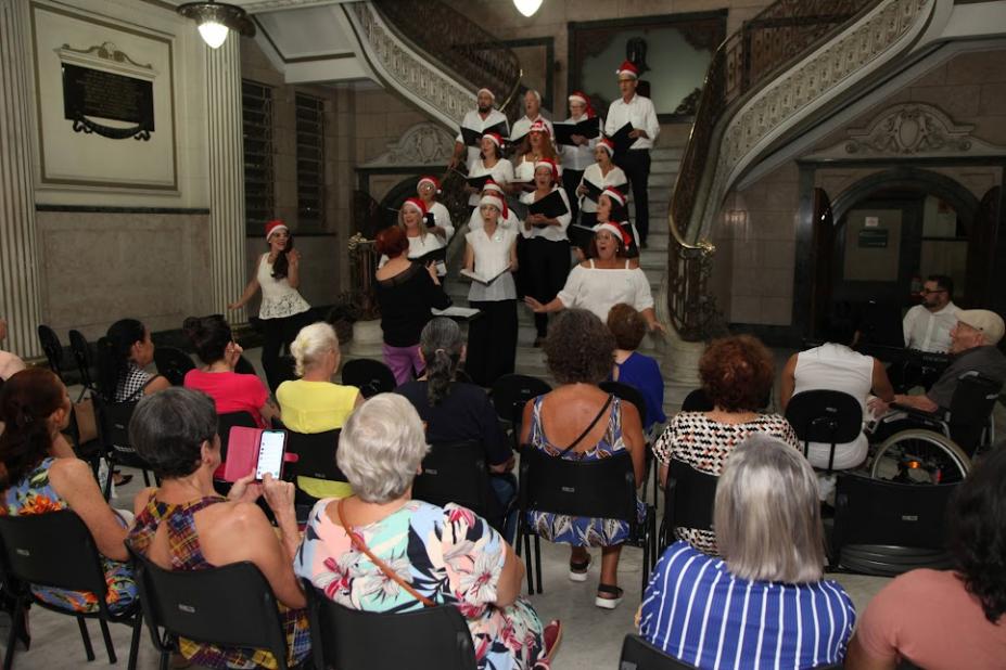 público assiste sentado apresentação de integrantes de coral na escadaria de saguão do paço. Os coralistas usam gorro de papai noel. #paratodosverem