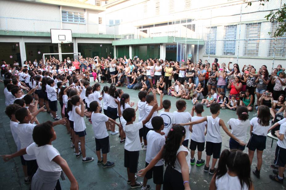 Crianças estão alinhadas em pátio de frente para as mães #pracegover 