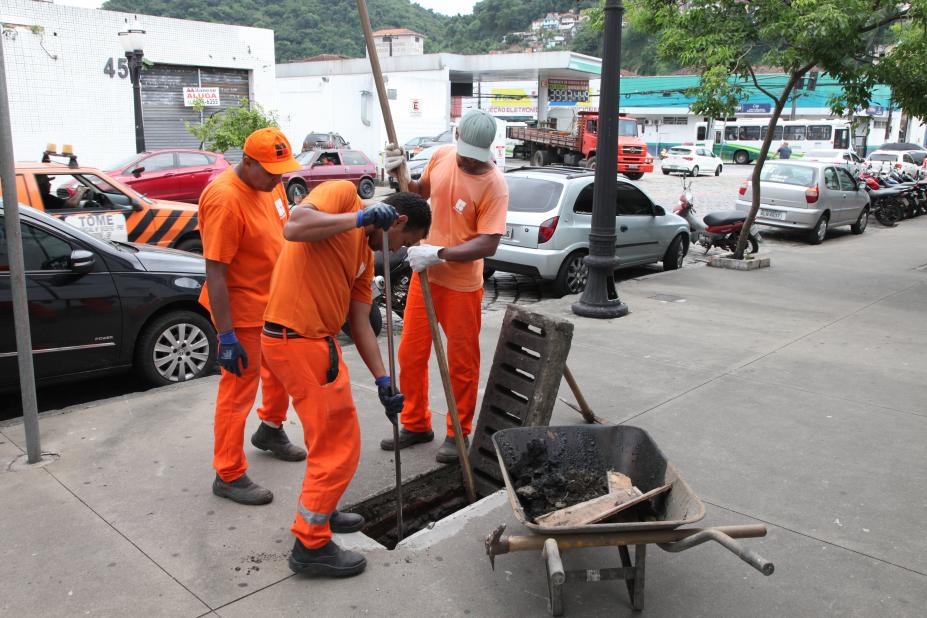 Funcionários uniformizados, com luvas e instrumentos de cabos longos para inserir na rede de drenagem fazem a limpeza em bueiro de calçada. #Pracegover