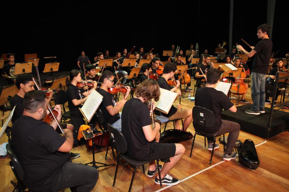 Jovens músicos estão sentados e tocando seus instrumentos. O maestro está à frente do grupo, sobre um degrau. #Pracegover