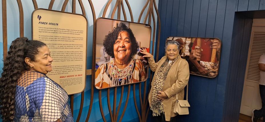 mulheres à frente de paineis de fotos sobre pesca. #paratodosverem