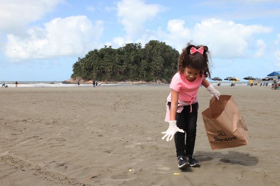criança usa luva para pegar um microlixo na faixa de areia. Ela segura um saco de papel para depositar o resíduo.#paratodosverem