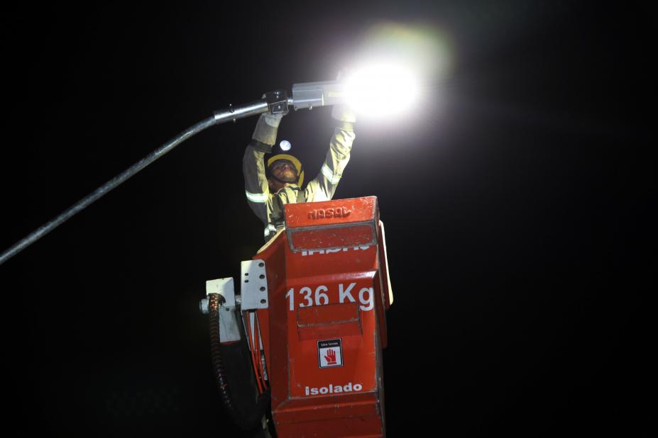 #pracegover Erguido dentro de cabine, homem de uniforme e luvas manuseia lâmpada de poste público à noite