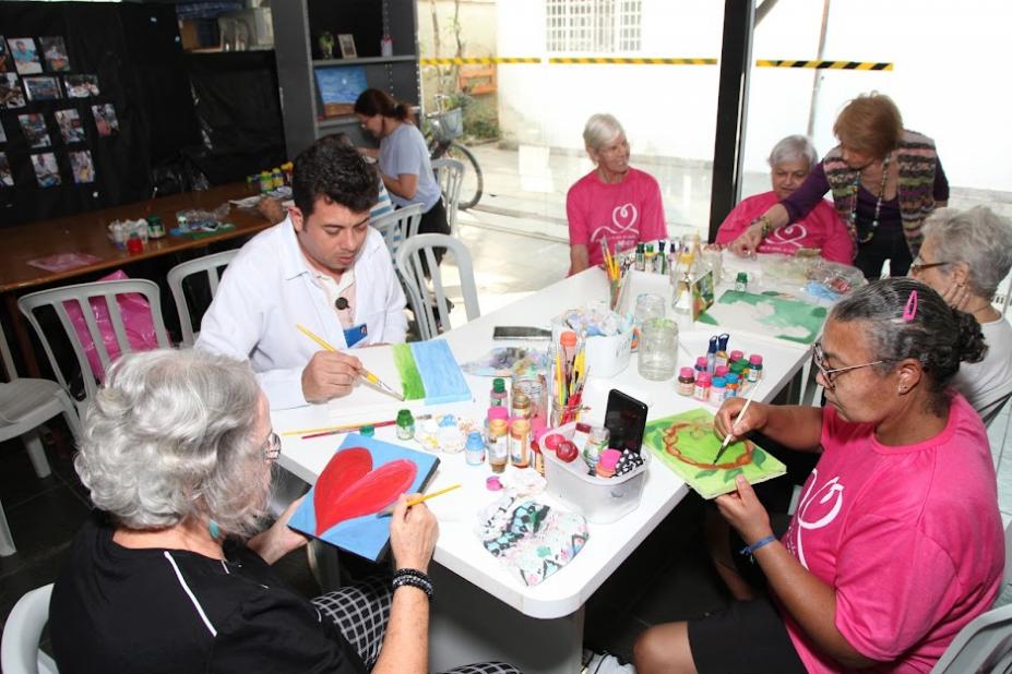 idosos estão sentados em torno de uma mesa pintando telas. A mesa está cheia de apetrechos como tintas e pinceis. #paratodosverem 