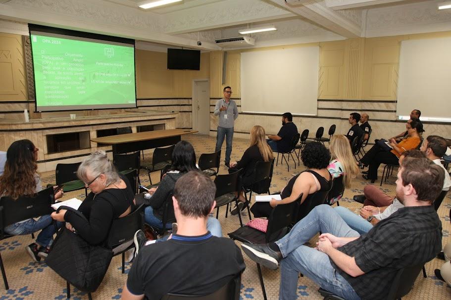 pessoas estão sentadas em auditório assistindo apresentação. Um homem fala a todos. #paratodosverem