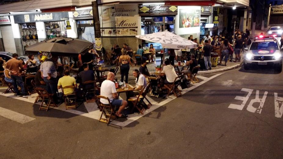 VÁRIAS PESSOAS SENTADAS EM MESAS DE BAR, COM CARRO DA POLÍCIA MILITAR PASSANDO AO LADO, BAR CHEIO #PARATODOSVEREM