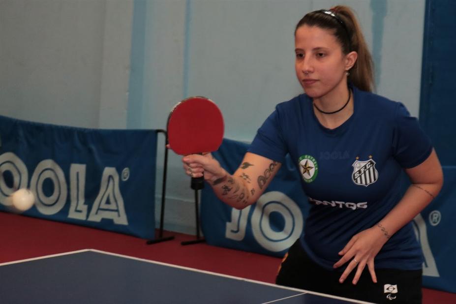 Jennyfer está segurando a raquete e a bola está no ar, à sua frente. #paratodosverem