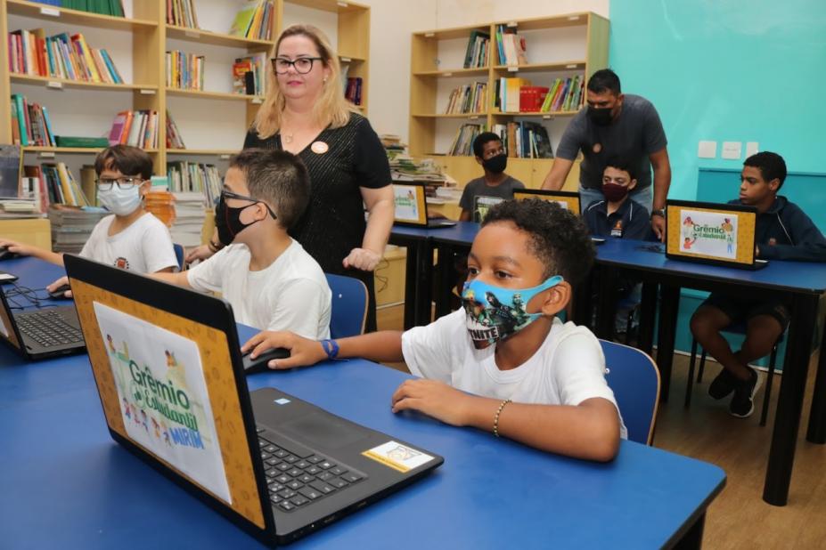 crianças estão sentadas em mesas diante de laptops. Em primeiro plano, o computador está aberto. Na tampa  se vê o nome do programa Grêmio Estudantil. #paratodosverem