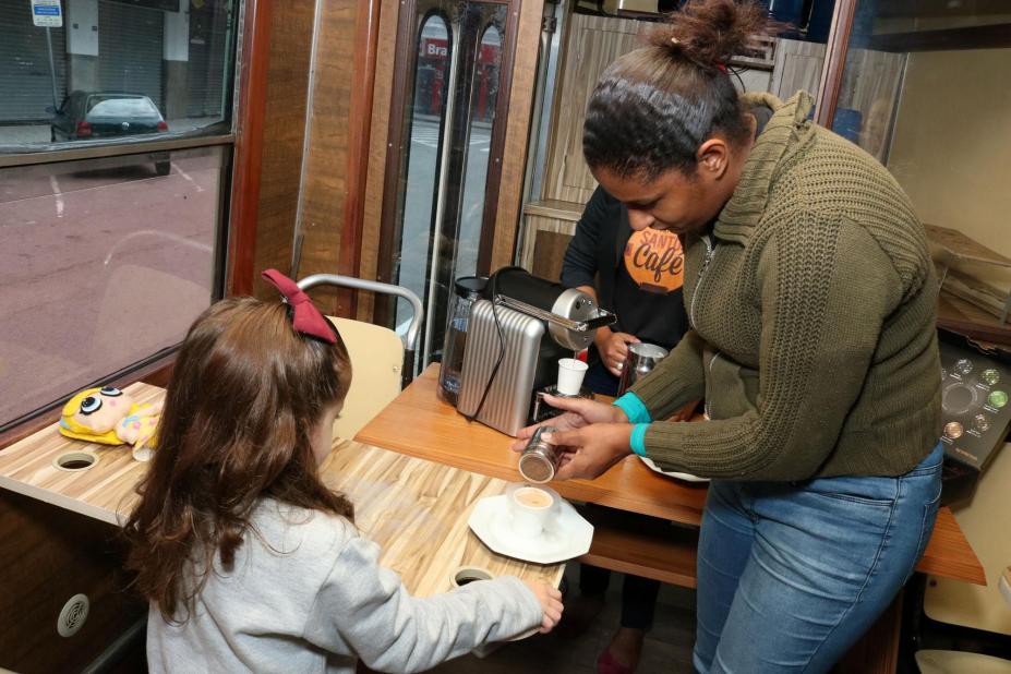 Mulher está em pé adiciona pó (canela ou chocolate) em xícara de café. Uma  menina, de costas para a imagem e com laço na cabeça, está segurando outra xícara de café. #Pracegover