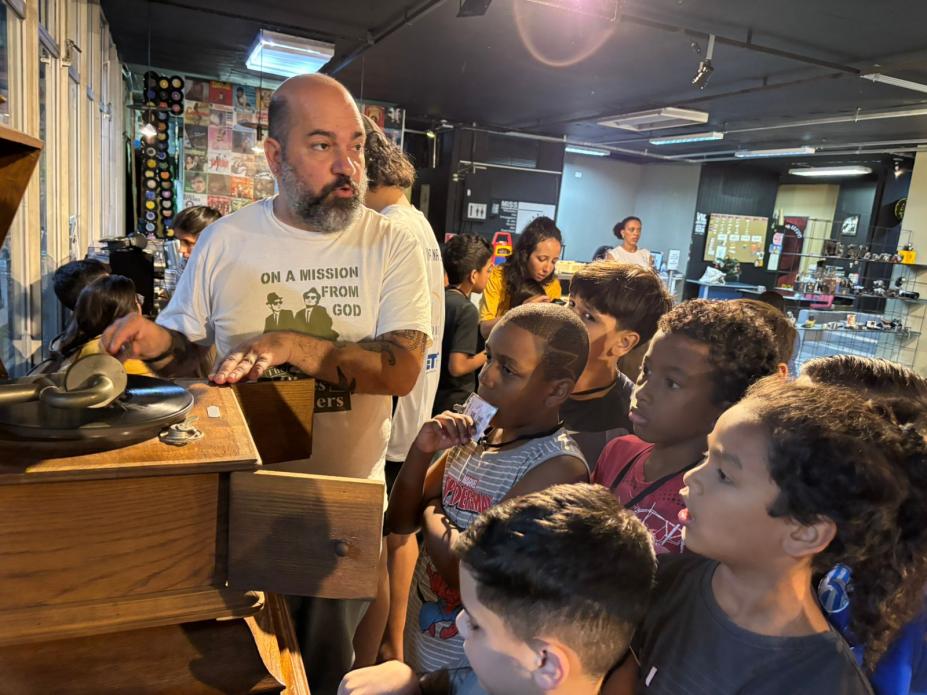 homem apresenta vitrola antiga de madeira para crianças reunidas em torno dele. #paratodosverem